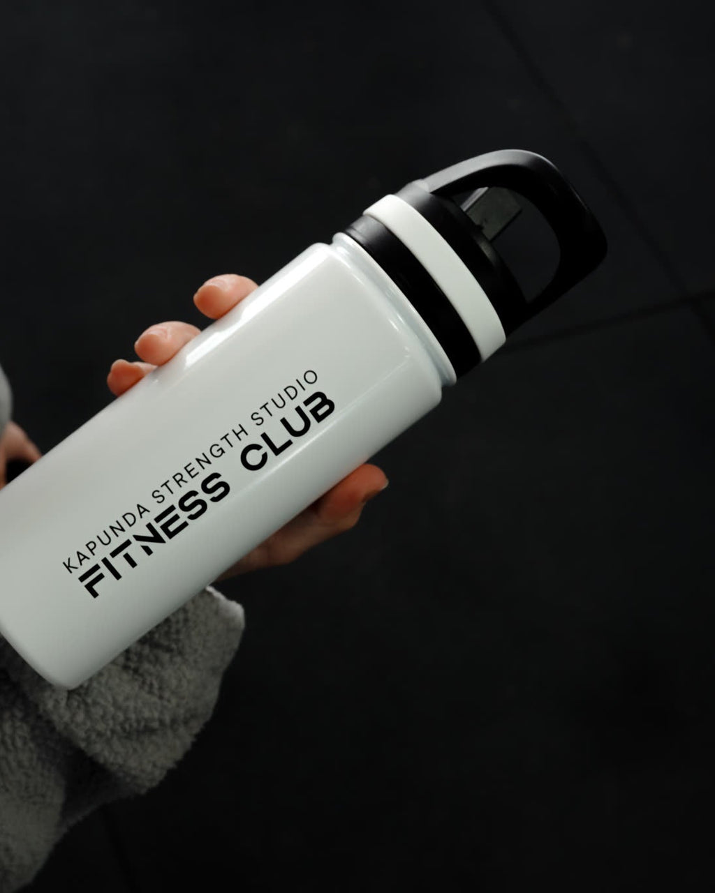 A person in a gray fleece jacket holds a white insulated water bottle with a black lid. The bottle is printed with the text 'KARUNISTRENGTHSTUDIO FITNESS CLUB'.