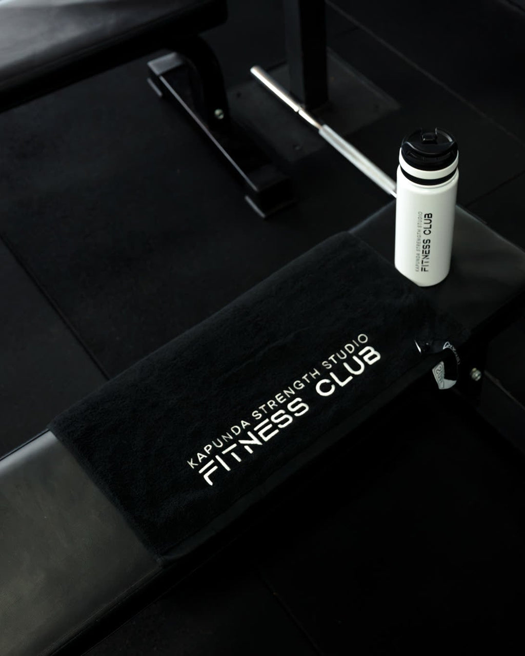 A black fitness bench with a black towel featuring white text that reads 'KAPUNA STRENGTH STUDIO FITNESS CLUB', with a white bottle placed on top, set in a dark gym environment.
