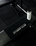 A black fitness bench with a black towel featuring white text that reads 'KAPUNA STRENGTH STUDIO FITNESS CLUB', with a white bottle placed on top, set in a dark gym environment.