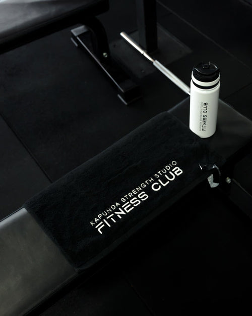A black fitness bench with a black towel featuring white text that reads 'KAPUNA STRENGTH STUDIO FITNESS CLUB', with a white bottle placed on top, set in a dark gym environment.