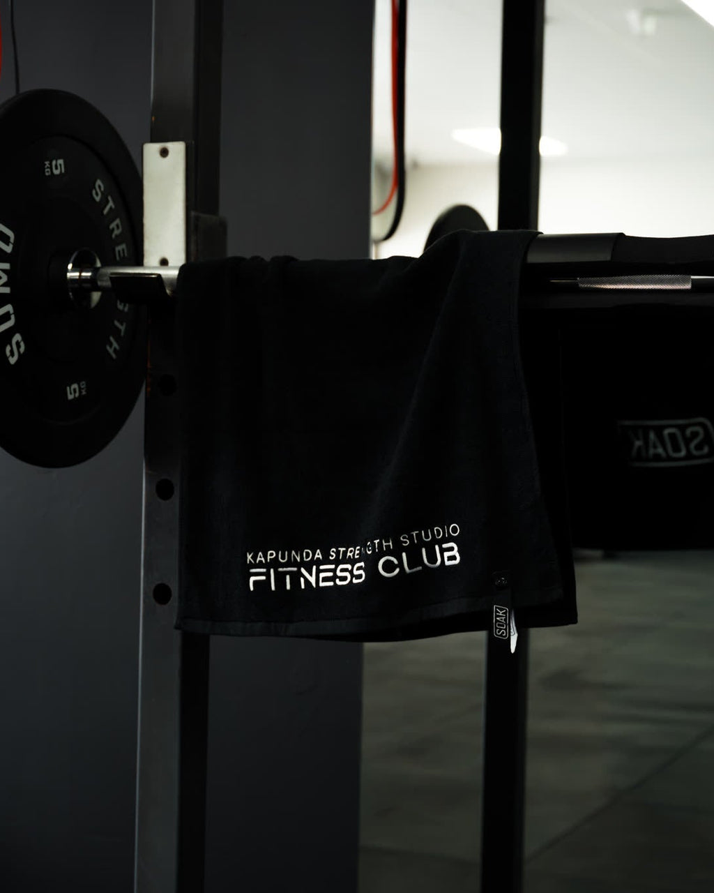A black gym towel with white text reading 'KARLUNA FITNESS STUDIO FITNESS CLUB' hanging from a barbell rack in a gym setting.