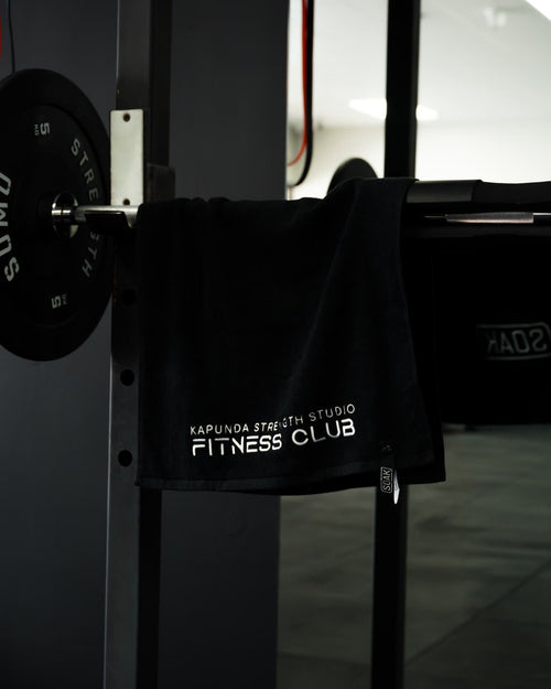 A black gym towel with white text reading 'KARLUNA FITNESS STUDIO FITNESS CLUB' hanging from a barbell rack in a gym setting.