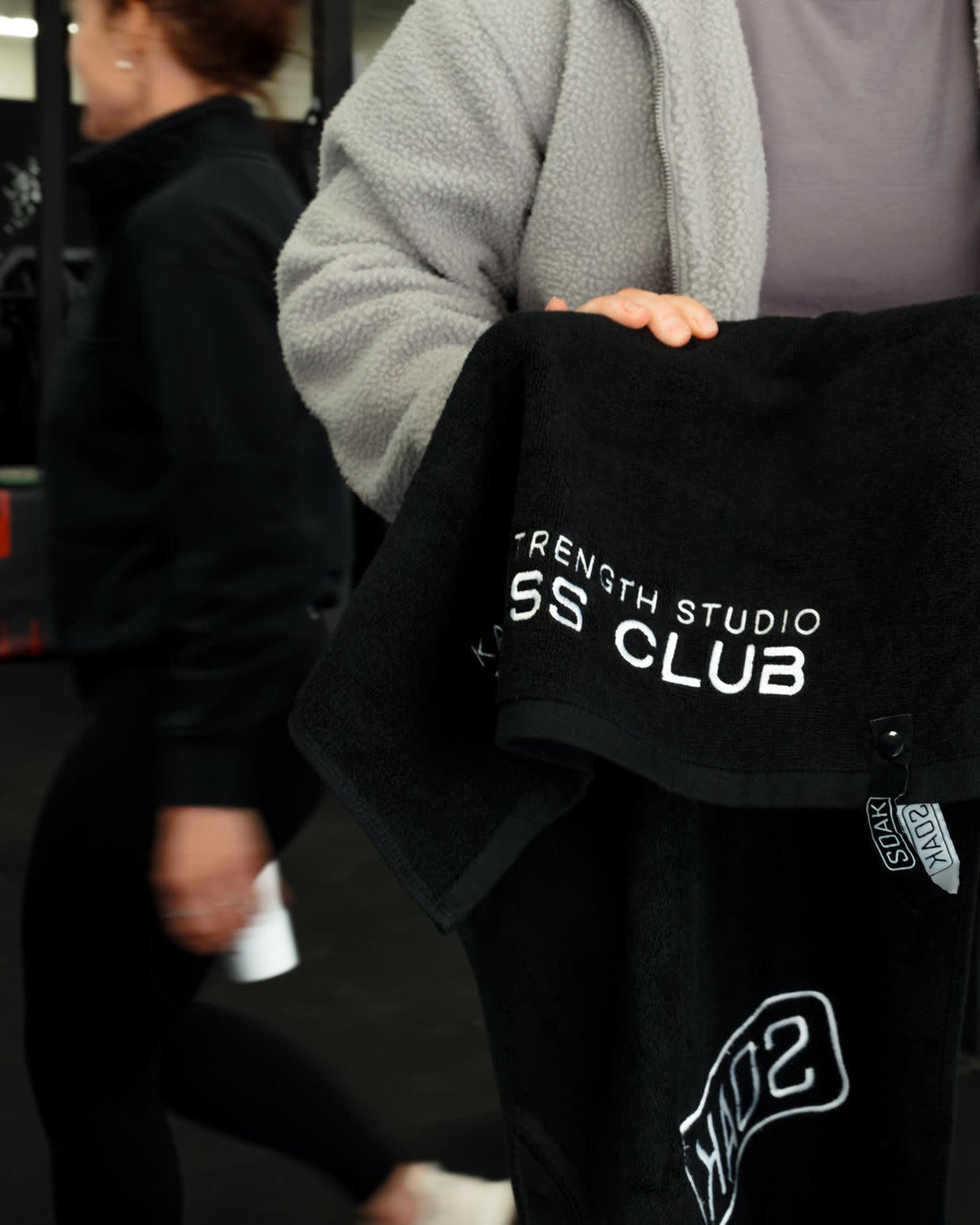 A black towel with white text reading 'SEITH STUDIO SS CLUB' is held up by a person in a gym setting. The person is wearing a gray zip-up hoodie and black sweatpants, and another person is visible in the background of the gym.
